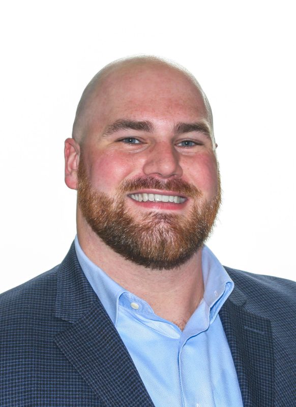 Clay Weissenborn '15, a bald man with a red beard, wearing a dark checkered suit jacket and a light blue dress shirt, smiling against a white background.