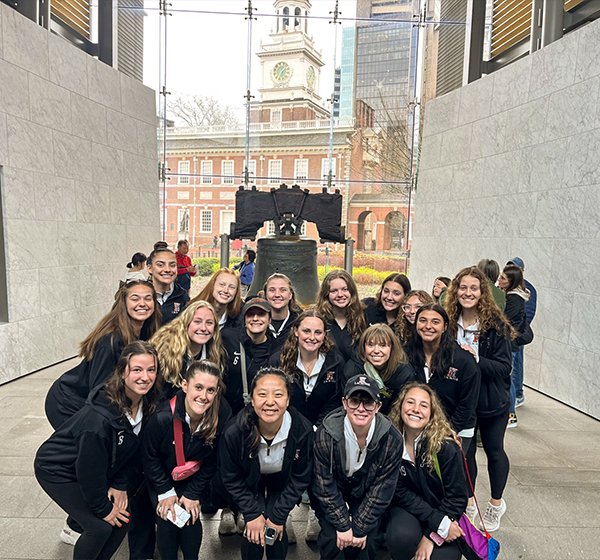 The Kalamazoo women's lacrosse team posing for a group photo in front of the Liberty Bell, with team members wearing black jackets featuring the Kalamazoo logo, some kneeling and others standing, in an outdoor setting with a historic building and clock tower in the background.