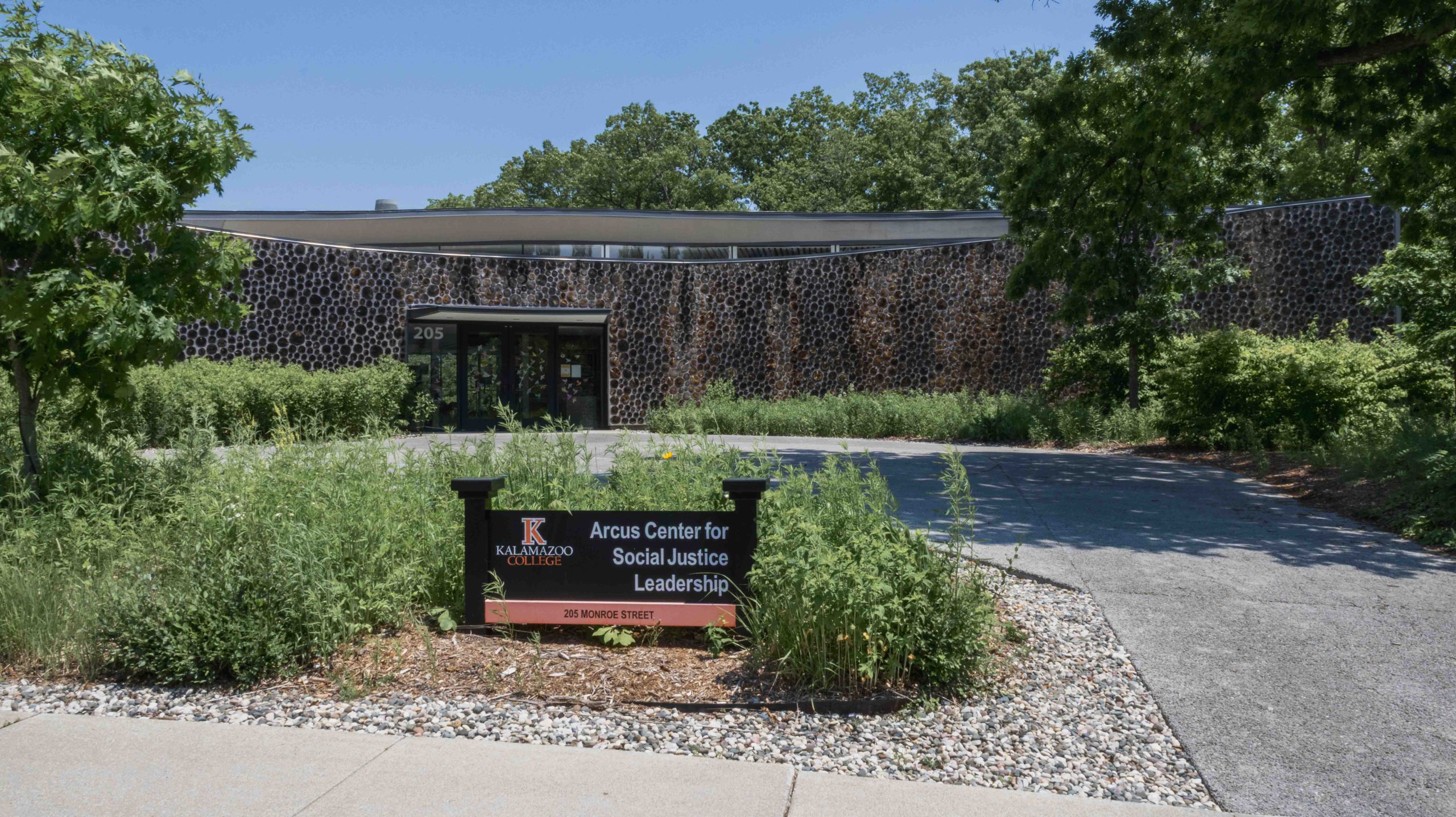 A modern building with a textured, circular-patterned exterior, identified as the Arcus Center for Social Justice Leadership at Kalamazoo College, located at 205 Monroe Street. The building is surrounded by lush greenery and trees under a clear blue sky, with a paved walkway leading to the entrance. The sign in the foreground clearly