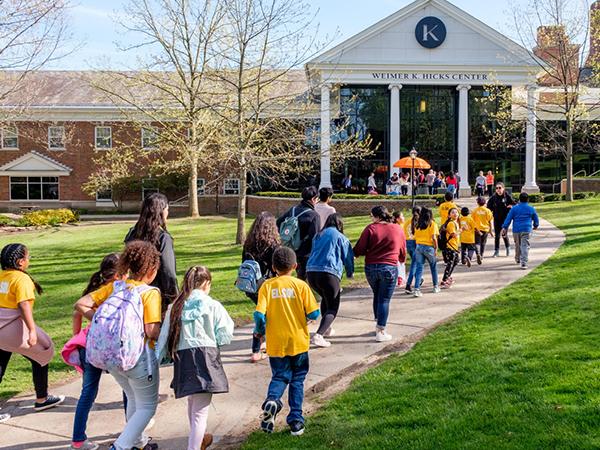A group of elementary school children, some wearing yellow t-shirts, walk along a paved path on a grassy lawn. They are on a field trip to Kalamazoo College and are being led by adults toward Hicks Center.