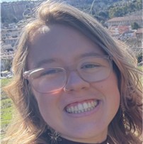 Maya Arau, a smiling woman with clear-framed glasses and light hair, posing outdoors with a scenic cityscape in the background.