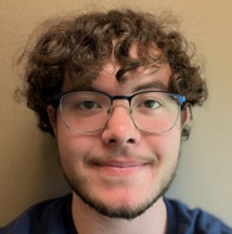 Cedric May, a smiling man with curly brown hair, glasses, and a light beard, set against a plain neutral background.