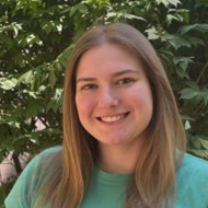 Sara Cate Finks, a smiling woman with straight light brown hair, wearing a teal shirt against a backdrop of green foliage.