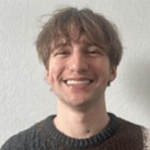 Luke Barnum, a smiling man with short brown hair and bangs, wearing a dark sweater against a plain white wall.