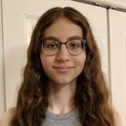 Juniper Pasternak, a person with long wavy brown hair and glasses, wearing a gray top against a white door background.