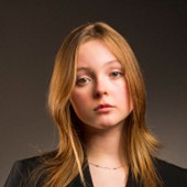 Thelma Hazen, a young woman with long strawberry blond hair wearing a dark jacket against a dark studio backdrop.