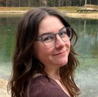 Elli Tiesworth, a smiling woman with dark wavy hair and glasses, posing outdoors with a body of water in the background.
