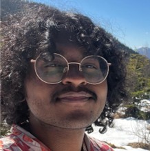 Newt Johnson, a smiling person with dark curly hair, round glasses, and a mustache, with a bright outdoor natural backdrop featuring a clear blue sky.
