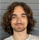 Charlie Doyle, a smiling man with shoulder-length curly brown hair and facial hair, wearing a white shirt against a gray horizontal-paneled wall background.