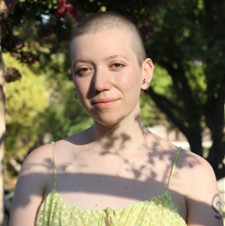 Alix Wilgoren, a person with a shaved head wearing a light green top, standing outdoors in soft dappled sunlight with trees in the background.