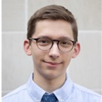 Benjamin Whitsett, a young man with short dark hair and round glasses, wearing a light blue dress shirt and blue tie against a light gray tiled background.