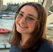 Isabelle Kastel, a smiling woman with straight brown hair and thin-framed glasses, posed against an outdoor waterfront backdrop with buildings in the background.