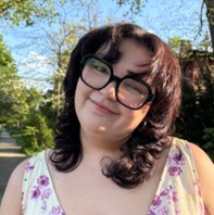 Estela Marquez, a smiling person with dark wavy hair and black-framed glasses, wearing a white floral top outdoors on a sunny day with trees in the background.