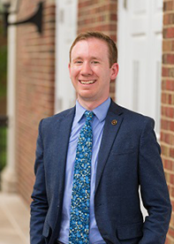 Andrew Grayson, a man with short reddish-brown hair, wearing a blue suit, light blue shirt, and a colorful patterned tie. He is standing in front of a brick wall with white doors and smiling at the camera.