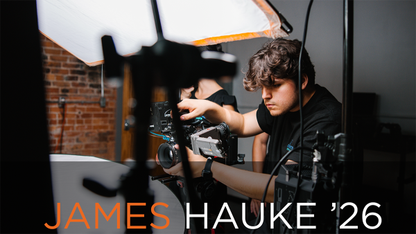 James Hauke '26 working with a camera setup at his internship. He is wearing a black shirt and adjusting the equipment, with a softbox light and brick wall in the background.