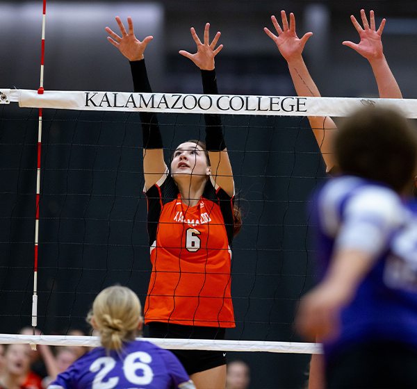 A volleyball player in an orange jersey with the number 6 and "Kalamazoo" written on it, jumping to block at the net during a match, with the "Kalamazoo College" banner visible. Opposing players in purple jerseys are also reaching up to block, and a crowd is in the background.