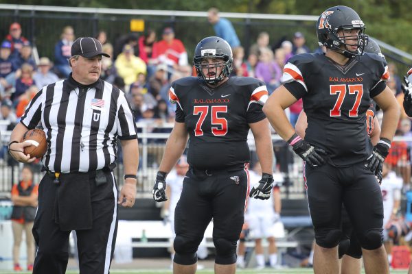 A football scene featuring Clay Weissenborn '15, a bald man with a red beard, wearing a black Kalamazoo College jersey with the number 75, standing on the field. He is flanked by a referee holding a football and another player in a jersey numbered 77, with a crowd and trees in the background.