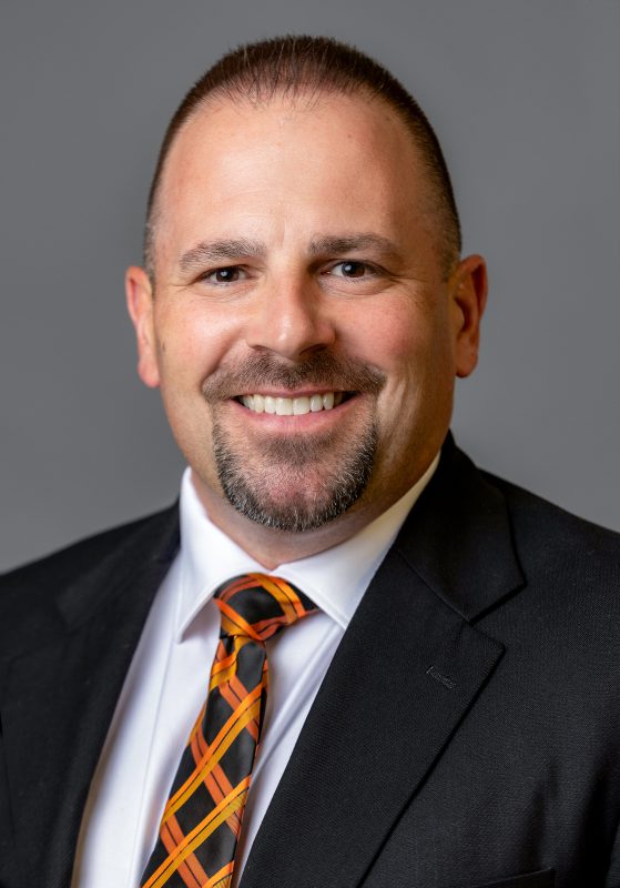 Jamie Zorbo, Director of Athletics, wearing a black suit, white dress shirt, and an orange and black patterned tie, smiling against a gray background.