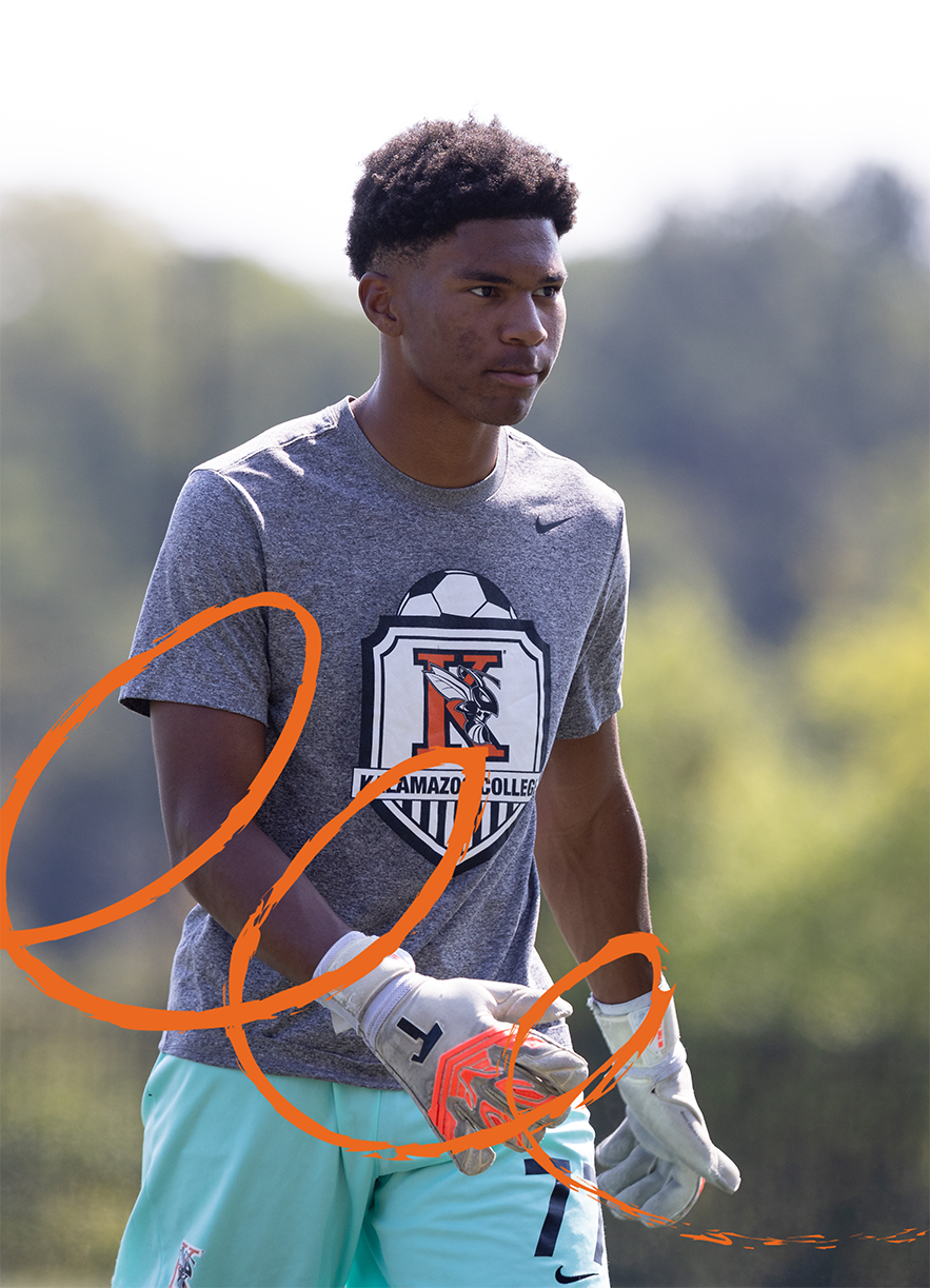 Darius Wright III ’27, in a Kalamazoo College soccer t-shirt and gloves stands outdoors. Orange circles graphic overlay. The background features a blurred green landscape.