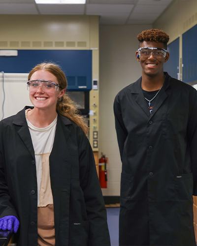 Two smiling students, posing in a science laboratory. Both are wearing black lab coats and clear safety goggles