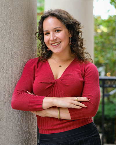 Olivia DiGiulio ’25 with curly brown hair leans against a large stone pillar outdoors. She is wearing a red long-sleeved shirt, black pinstriped pants, and gold jewelry.
