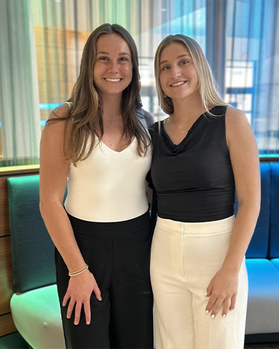 Savannah Chapie ’27 and Jillian Smith ’27 standing side-by-side indoors. Savannah (left) has long brown hair and wears a white tank top with black pants. Jillian (right) has blonde hair and wears a black sleeveless top with white pants.