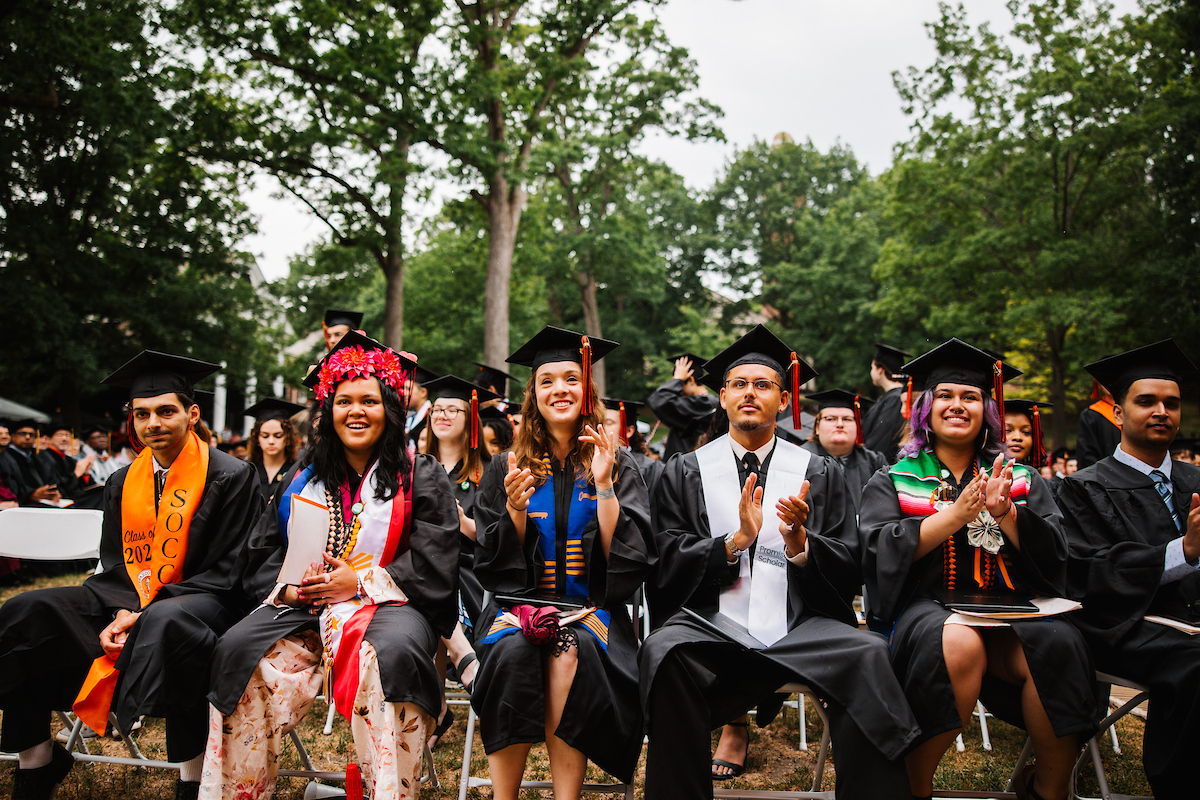 A diverse group of Kalamazoo College graduates in black caps and gowns, featuring colorful stoles and floral headwear, smiling and clapping during an outdoor graduation ceremony.