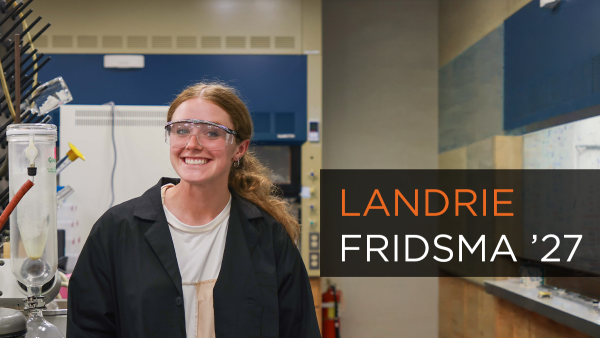 Landrie Fridsma ’27, is pictured in a science laboratory. She is wearing a black lab coat over a white shirt and clear protective safety goggles. To her right, a large black text box with white and orange lettering identifies her as LANDRIE FRIDSMA ’26.
