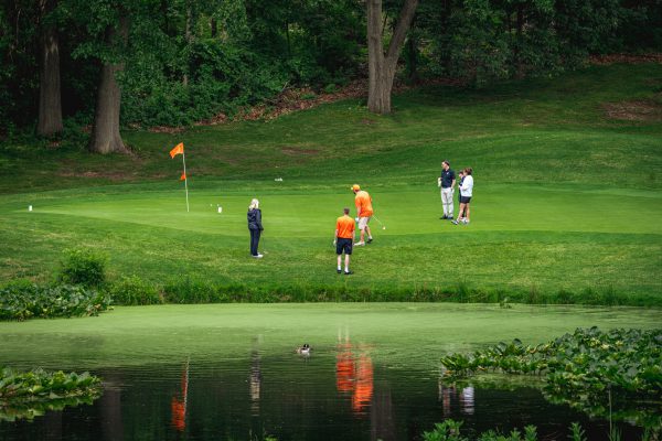 Wide view of a golf course green with an orange flag, a water hazard in the foreground, and several golfers lining up putts near the hole.