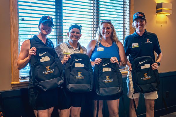 Four women smiling indoors as they pose together, each holding up a black Nike backpack featuring the Kalamazoo Athletics logo.