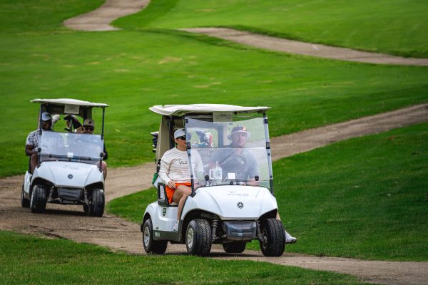 Two golf carts carrying participants driving along a paved cart path on a green golf course fairway.