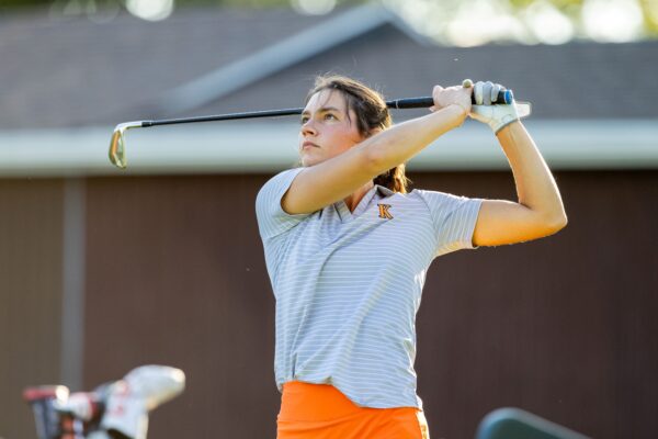 A female student-athlete in a grey striped Kalamazoo College polo and orange skirt watching her golf shot after finishing a swing.