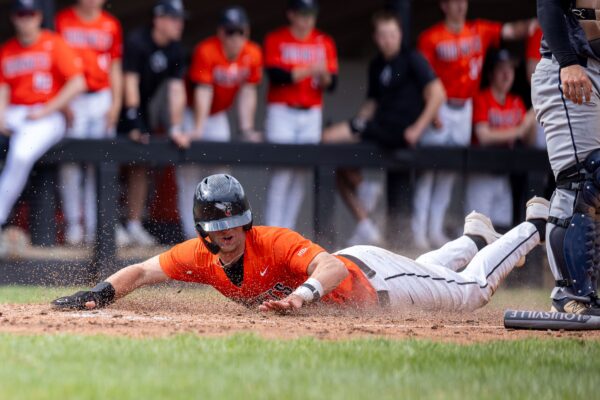 A baseball player wearing an orange jersey sliding headfirst into a base, kicking up dirt as teammates watch from the dugout.