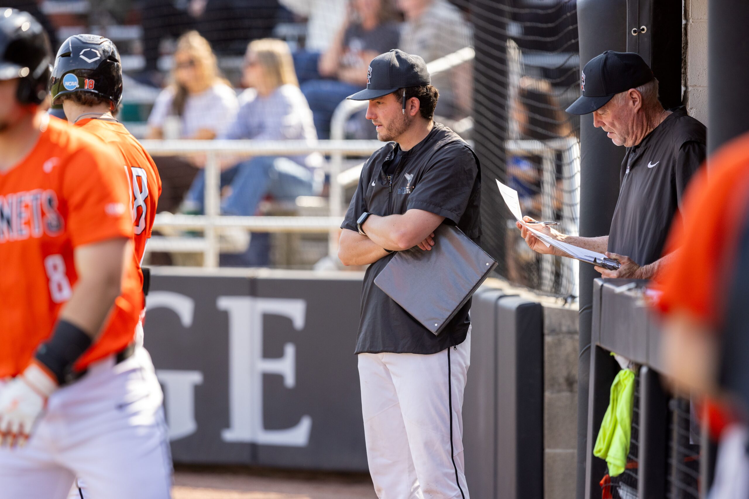 Two Kalamazoo College baseball coaches standing near the dugout during a game, with one coach holding a binder and arms crossed while another reviews papers on a clipboard.