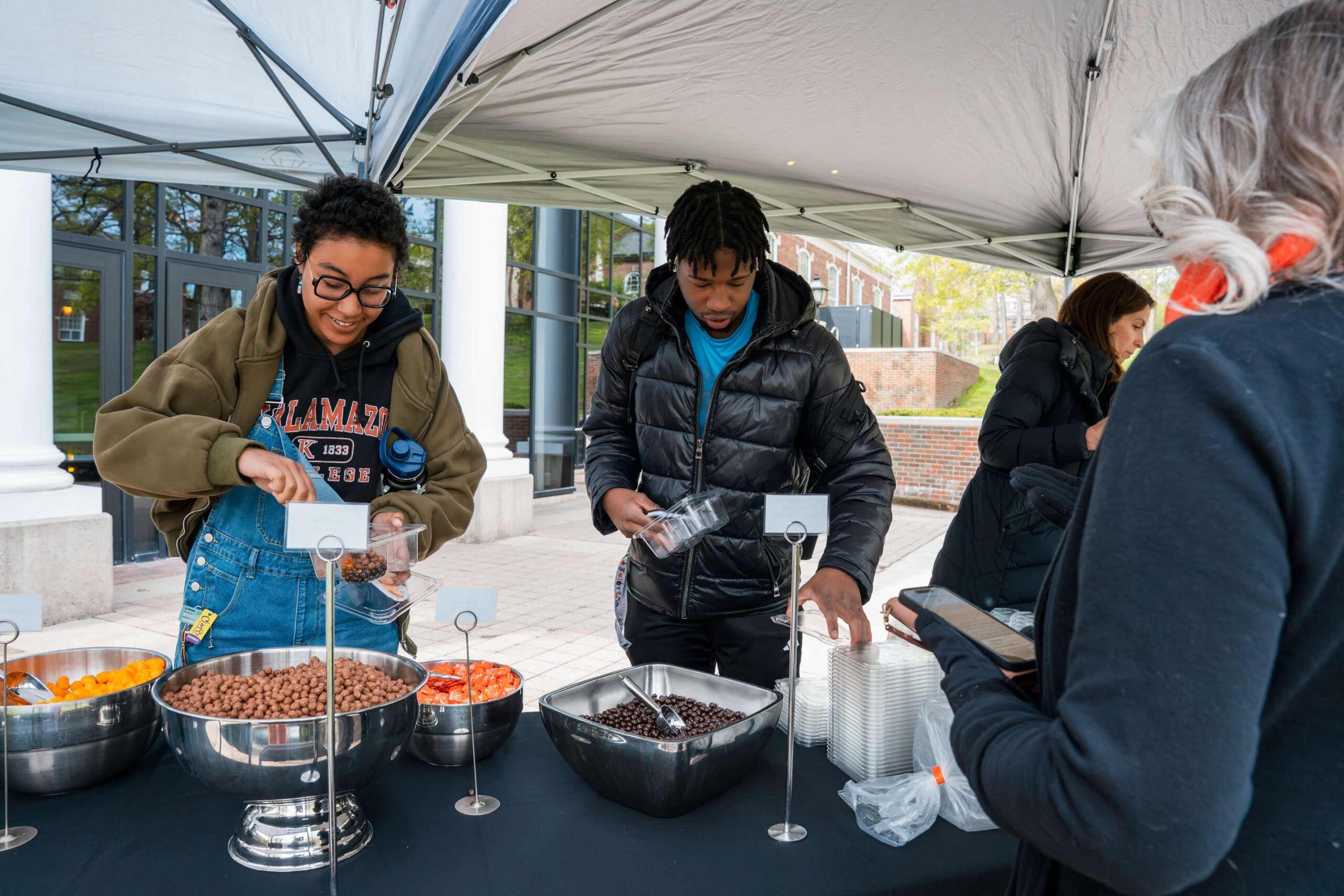 Two students scooping snacks out of metal bowls with two staff members assisting. 