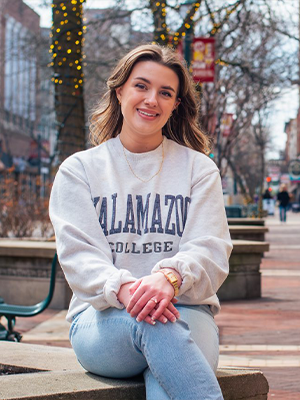 Ariadne Markou ’25 sits on a low stone wall in an outdoor urban setting, wearing a light gray sweatshirt with "Kalamazoo College" printed on it, light blue jeans, and a watch. The background features leafless trees wrapped in string lights, brick buildings, and a few people walking along a pathway.