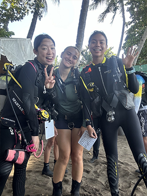 Brooke Dolhay ’25 stands on a sandy beach with two other individuals, all wearing scuba diving gear including wetsuits and tanks. One person makes a peace sign, another waves, and they are surrounded by palm trees and a tent in the background.