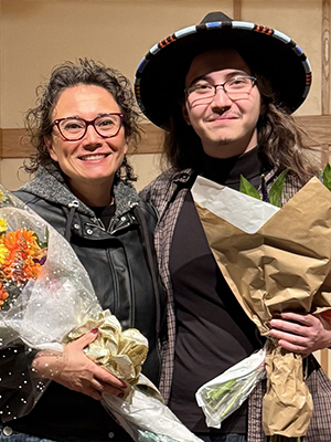 Davis Henderson ’25 and his mother Holly stand side by side, each holding a bouquet of flowers wrapped in paper. Davis wears a wide-brimmed hat and a dark shirt, while Holly is dressed in a black jacket and glasses. The background features a wooden wall and a warm indoor setting.