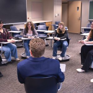 Students in a classroom sit in a semicircle around their professor