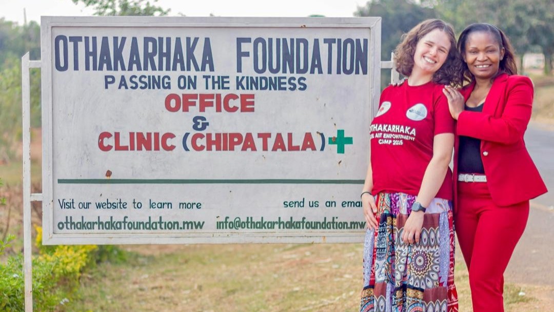 Two women in front of a sign that says Othakarhaka Foundation, Passing on the Kindness