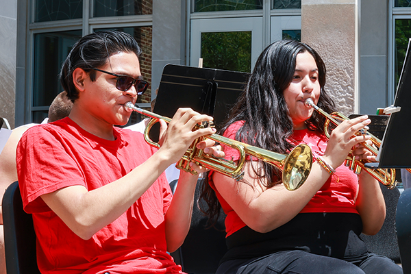 Two students wearing red shirts are playing trumpets while seated and reading sheet music from stands, with a building and windows in the background.