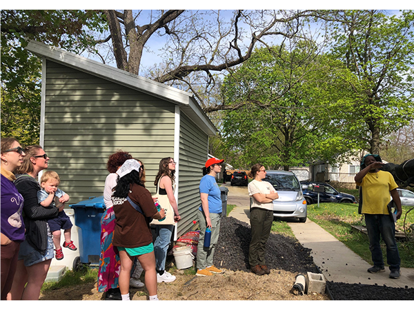 A group of Kalamazoo students stand on a dirt and gravel patch next to a building. Ben Brown in a yellow shirt gestures while speaking to the students, who are listening attentively. This photo documents a gathering for the Kalamazoo College Just Food Collective, a student-led, sustainable-food systems program.