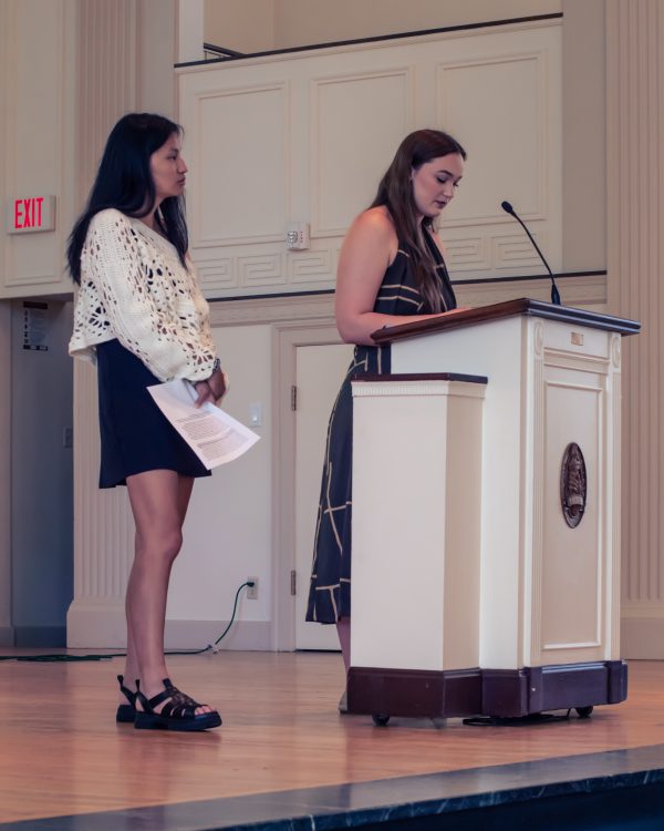 Two Kalamazoo College students participating in a Community Reflection event, with one standing at a podium delivering a speech and the other holding papers nearby.